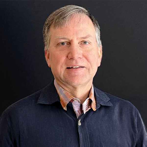 Man in a collared shirt and dark jacket standing against a plain black background.