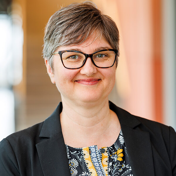 Smiling person with short gray hair, glasses, and a black blazer.
