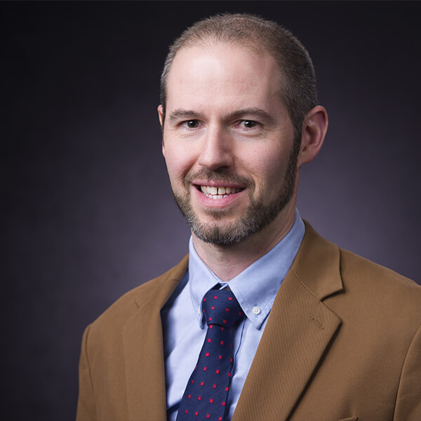 Man in brown suit, blue shirt, and tie, smiling in front of a dark background.