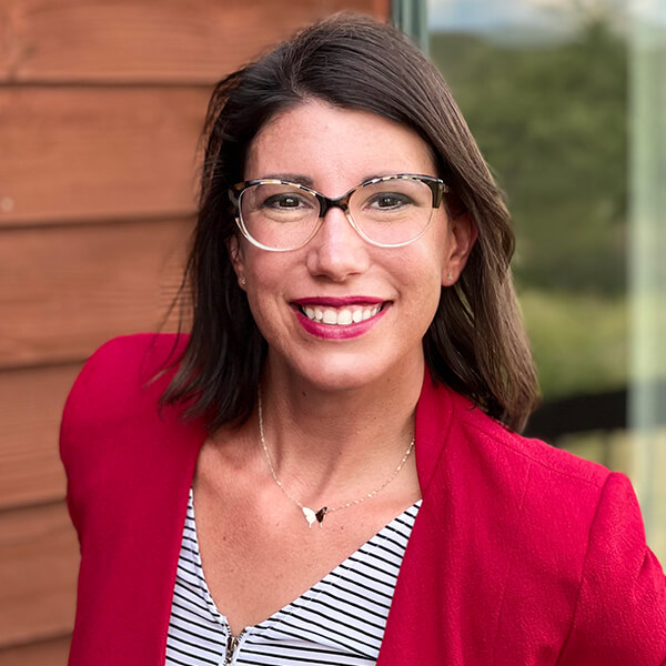 Woman in glasses and red blazer smiling outside, wooden wall and greenery in background.